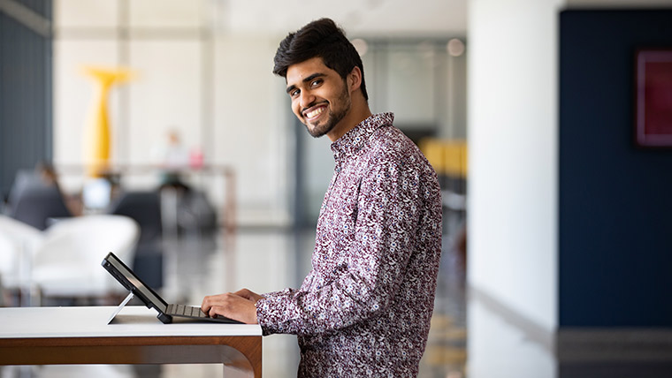 A student smiles toward the camera while working on his laptop.